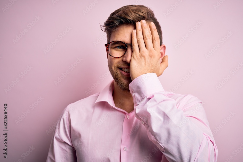 Young handsome blond man with beard and blue eyes wearing pink shirt and glasses covering one eye with hand, confident smile on face and surprise emotion. Young handsome blond man with beard and blue eyes wearing pink shirt and glasses covering one eye with hand, confident smile on face and surprise emotion.