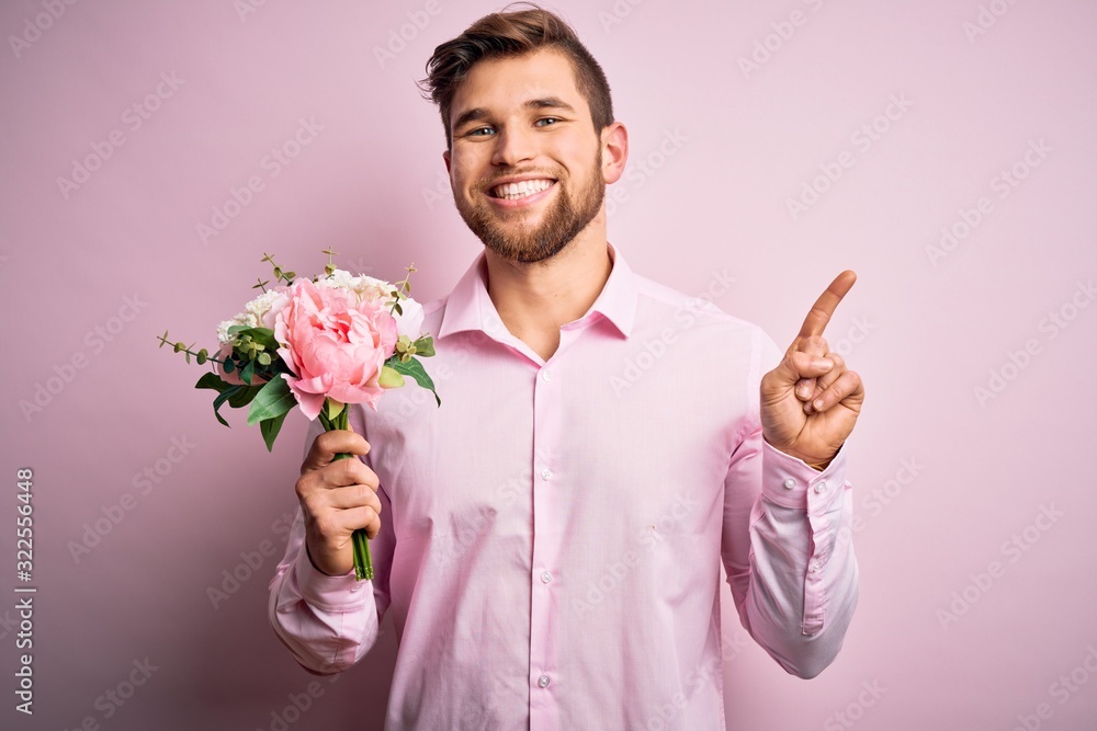 Young blond romantic man with beard and blue eyes holding beautiful bouquet of flowers surprised with an idea or question pointing finger with happy face, number one Young blond romantic man with beard and blue eyes holding beautiful bouquet of flowers surprised with an idea or question pointing finger with happy face, number one