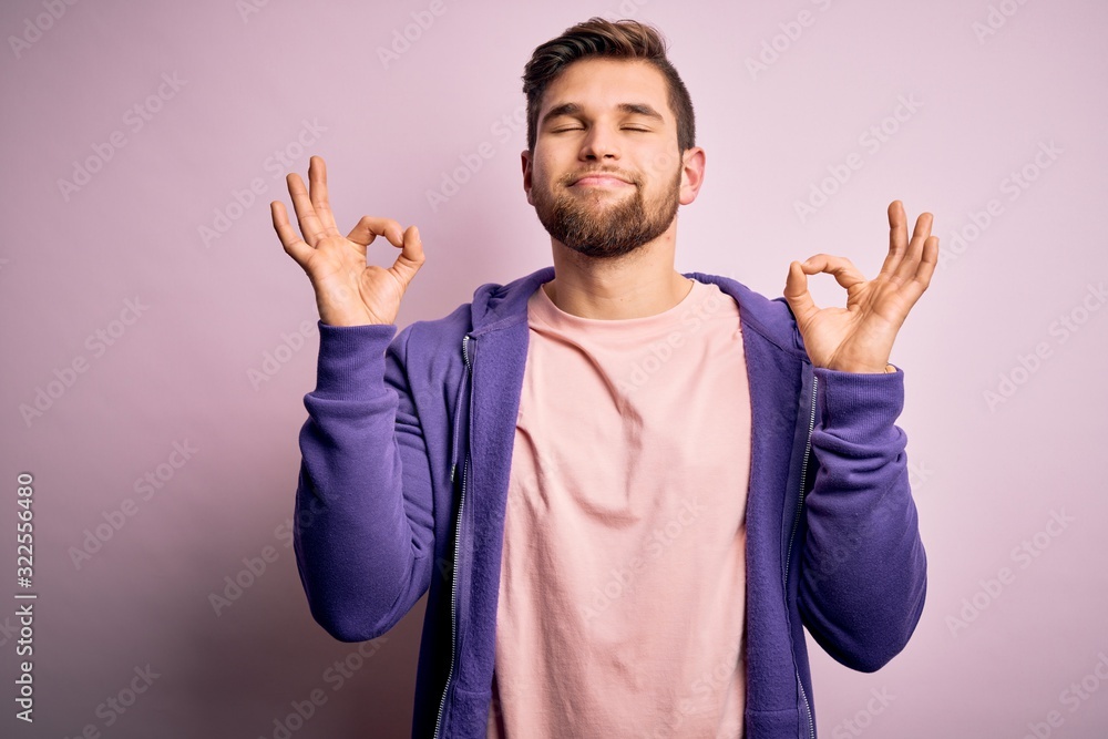 Fototapeta premium Young blond man with beard and blue eyes wearing purple sweatshirt over pink background relax and smiling with eyes closed doing meditation gesture with fingers. Yoga concept.