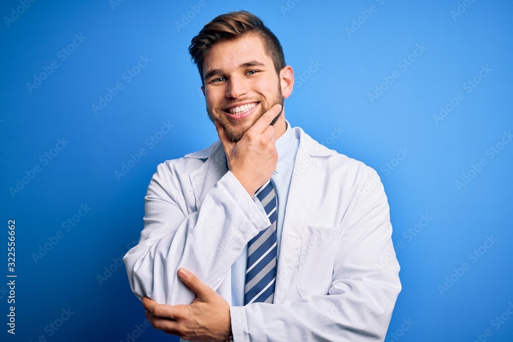 Young blond therapist man with beard and blue eyes wearing coat and tie over background looking confident at the camera smiling with crossed arms and hand raised on chin. Thinking positive. Young blond therapist man with beard and blue eyes wearing coat and tie over background looking confident at the camera smiling with crossed arms and hand raised on chin. Thinking positive.