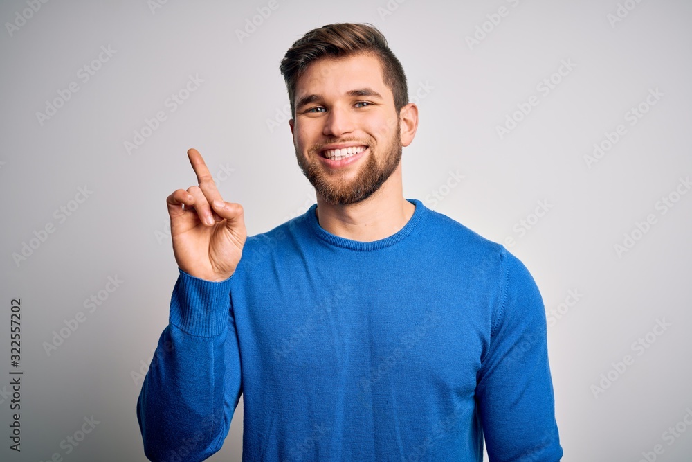 Young handsome blond man with beard and blue eyes wearing casual sweater showing and pointing up with finger number one while smiling confident and happy. Young handsome blond man with beard and blue eyes wearing casual sweater showing and pointing up with finger number one while smiling confident and happy.