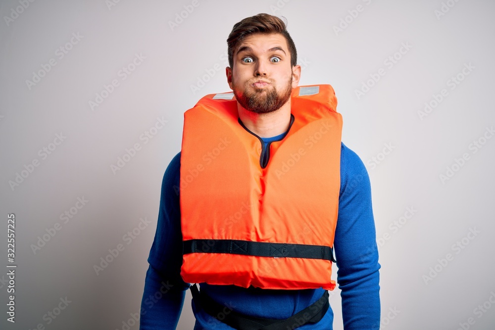 Young blond tourist man with beard and blue eyes wearing lifejacket over white background puffing cheeks with funny face. Mouth inflated with air, crazy expression. Young blond tourist man with beard and blue eyes wearing lifejacket over white background puffing cheeks with funny face. Mouth inflated with air, crazy expression.