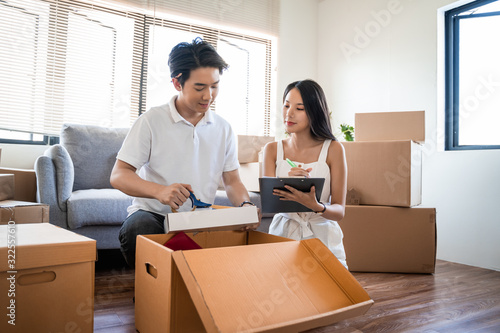 Young beautiful asian couple in love moving to new home, sitting on the floor very happy and cheerful for new apartment around cardboard boxes and holding cardboard boxes while moving home