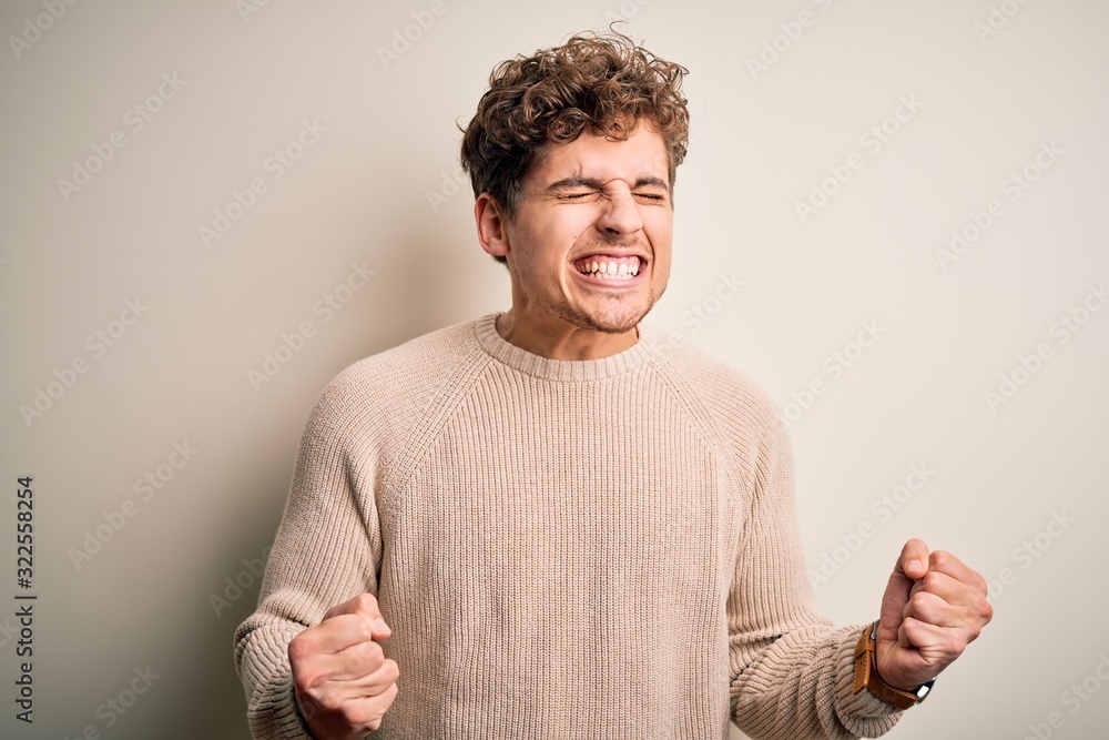 Young blond handsome man with curly hair wearing casual sweater over white background excited for success with arms raised and eyes closed celebrating victory smiling. Winner concept. Young blond handsome man with curly hair wearing casual sweater over white background excited for success with arms raised and eyes closed celebrating victory smiling. Winner concept.