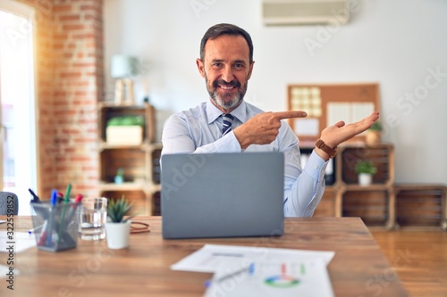 Middle age handsome businessman wearing tie sitting using laptop at the office amazed and smiling to the camera while presenting with hand and pointing with finger.