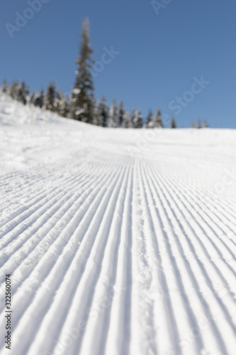 Fresh groomed snow on ski slope at ski resort on a sunny winter day. snow groomer tracks on a mountain ski piste. snowy spruces in the background © Lukasz