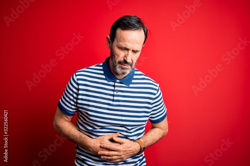 Middle age hoary man wearing casual striped polo standing over isolated red background with hand on stomach because indigestion, painful illness feeling unwell. Ache concept.