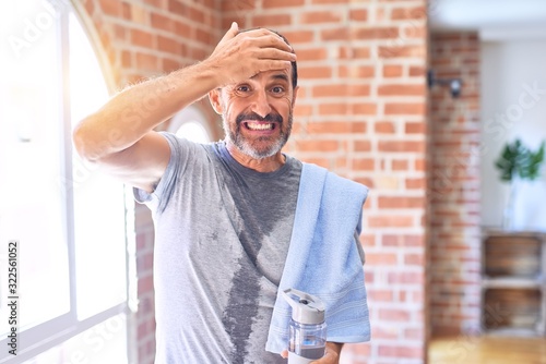 Canvas Print Middle age handsome sportman sweaty holding bottle of water and towel after exercise at gym stressed with hand on head, shocked with shame and surprise face, angry and frustrated