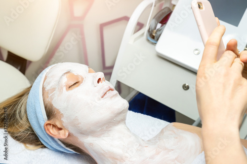 Young adult woman lying on bed at beauty spa salon with applied facial cosmetic mask making phone selfie shot for social networks. Influencer making online vlog stream with skin care product review