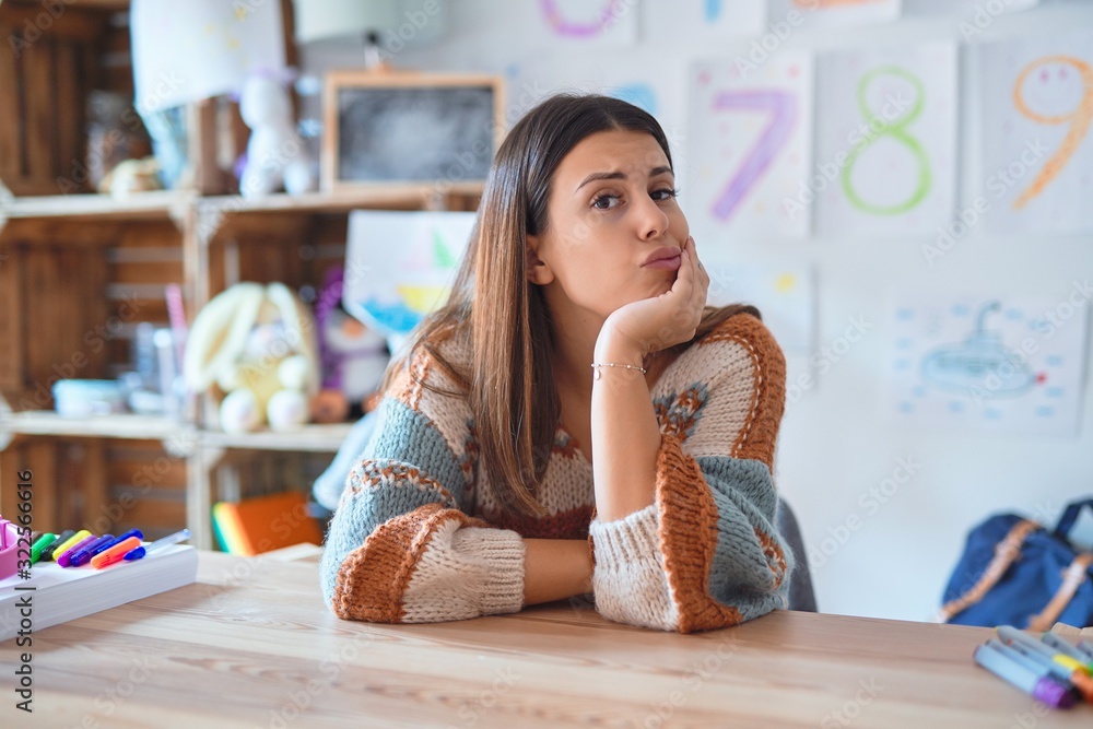 Young beautiful teacher woman wearing sweater and glasses sitting on desk at kindergarten thinking looking tired and bored with depression problems with crossed arms.