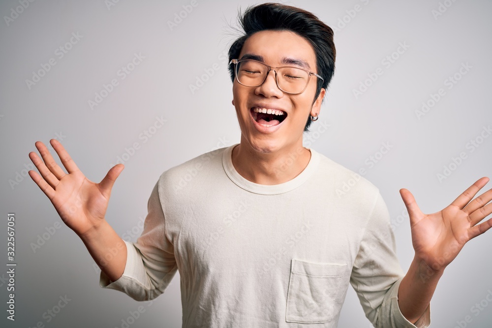Young handsome chinese man wearing casual t-shirt and glasses over white background celebrating mad and crazy for success with arms raised and closed eyes screaming excited. Winner concept Young handsome chinese man wearing casual t-shirt and glasses over white background celebrating mad and crazy for success with arms raised and closed eyes screaming excited. Winner concept