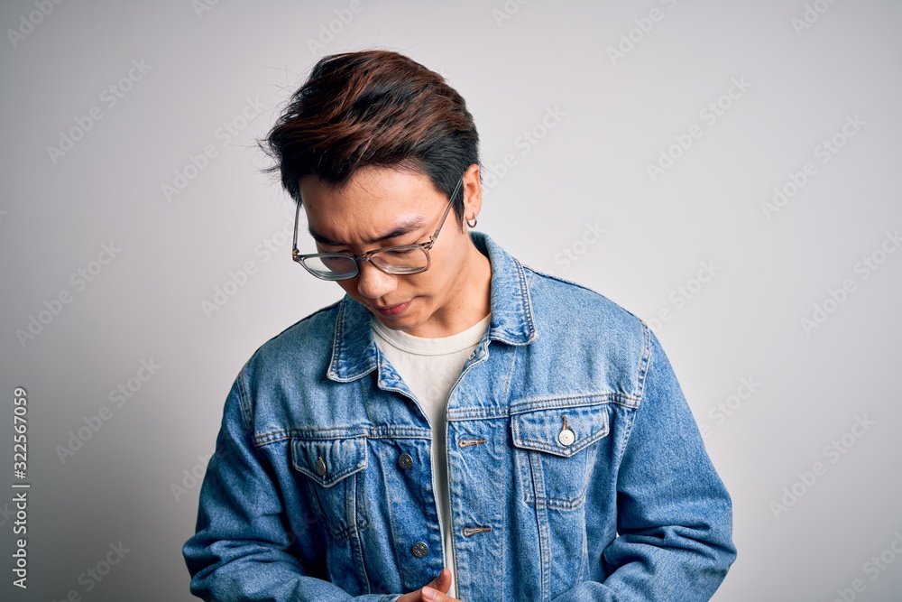 Young handsome chinese man wearing denim jacket and glasses over white background with hand on stomach because indigestion, painful illness feeling unwell. Ache concept. Young handsome chinese man wearing denim jacket and glasses over white background with hand on stomach because indigestion, painful illness feeling unwell. Ache concept.
