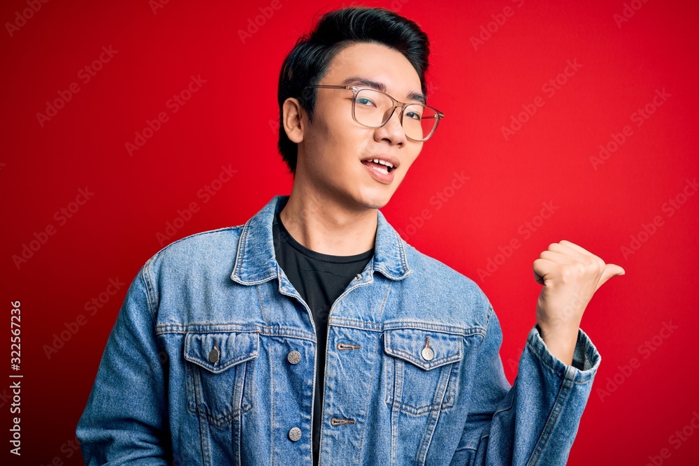Young handsome chinese man wearing denim jacket and glasses over red background smiling with happy face looking and pointing to the side with thumb up. Young handsome chinese man wearing denim jacket and glasses over red background smiling with happy face looking and pointing to the side with thumb up.