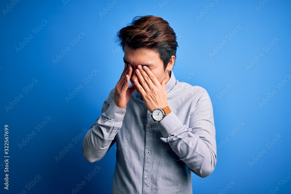 Young handsome chinese man wearing casual shirt standing over isolated blue background rubbing eyes for fatigue and headache, sleepy and tired expression. Vision problem Young handsome chinese man wearing casual shirt standing over isolated blue background rubbing eyes for fatigue and headache, sleepy and tired expression. Vision problem