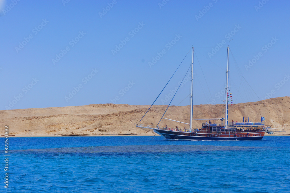 Fototapeta premium A sailing ship with masts and people on a blue sea against a yellow sandy shore