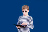 Blonde child boy with big glasses playing with tablet pc on blue background. Education concept.