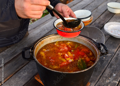 A man, a cook, pours a tomato and pepper soup into a red plate from a large saucepan. Healthy food, lunch in nature