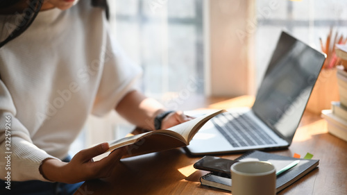 Cropped image of young beautiful woman holding/reading a books in her hands in front a computer laptop, coffee cup and smartphone at the modern wooden table with comfortable living room as background.