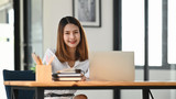 Photo of young creative woman in white comfortable shirt working in front computer laptop at the modern wooden table with office wall glass as background.