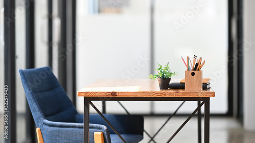 Side shot of armchair setting together with wooden table and office accessories putting on it with office glass wall as background. Comfortable working space concept. Vintage decoration style.