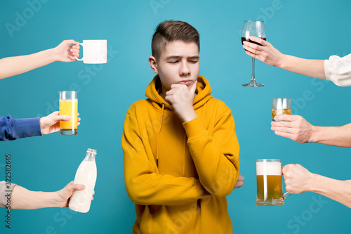 A young man in a yellow sweatshirt on a blue background looks doubtfully at an imaginary object to his left, holding his hand on his chin photo