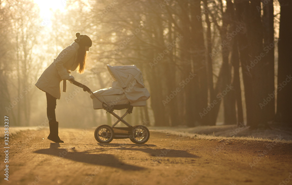 Silhouette of young woman on sunny footpath and looking at child in baby stroller. Sunny winter park, childcare and love Silhouette of young woman on sunny footpath and looking at child in baby stroller. Sunny winter park, childcare and love