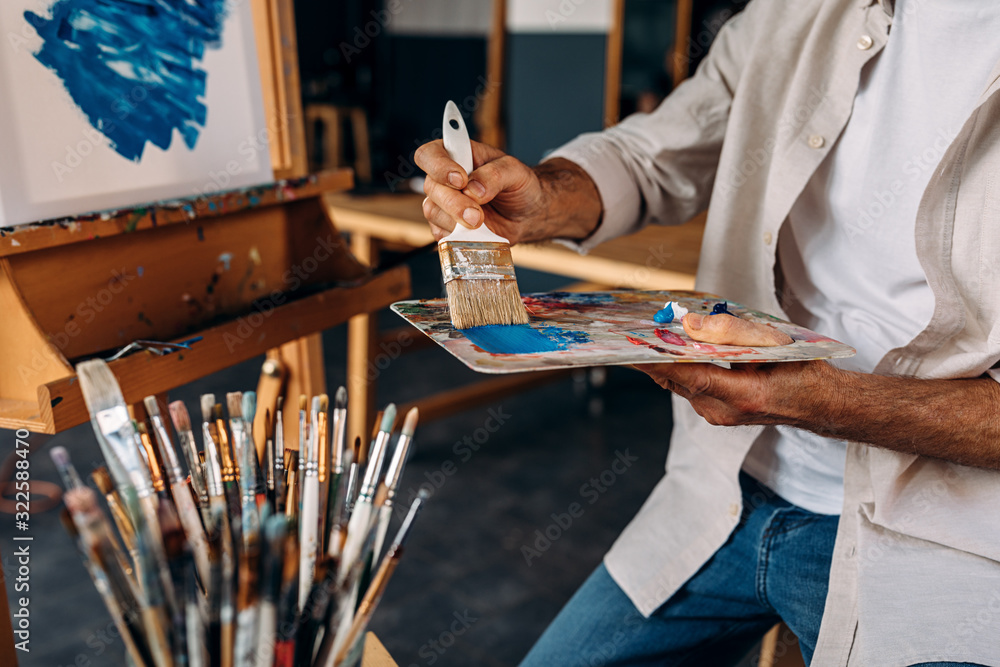 Fototapeta premium Cropped shot of artist sitting in front of an easel and dipping a large paintbrush into a colorful palette