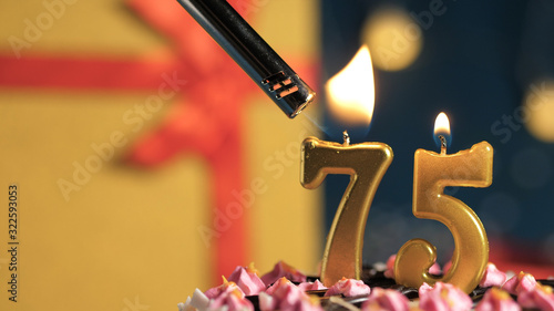 Birthday cake number 75 golden candles burning by lighter, background gift yellow box tied up with red ribbon. Close-up photo