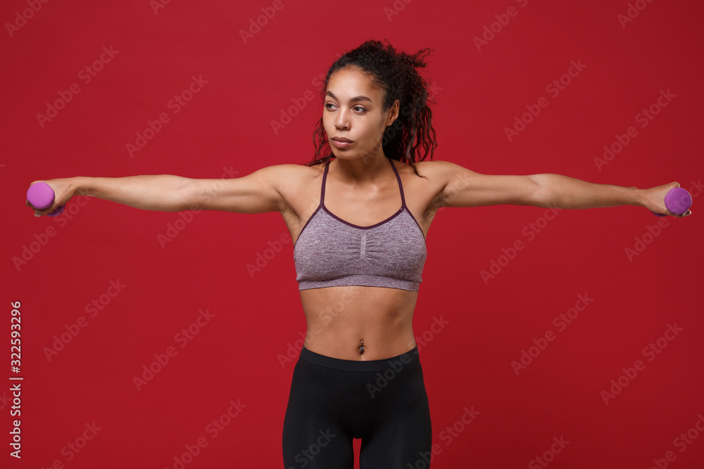 Beautiful young african american sports fitness woman in sportswear work out isolated on red background. Sport exercises healthy lifestyle concept. Spreading hands make exercise for arm with dumbbell.