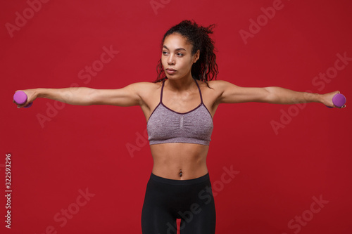 Beautiful young african american sports fitness woman in sportswear work out isolated on red background. Sport exercises healthy lifestyle concept. Spreading hands make exercise for arm with dumbbell.