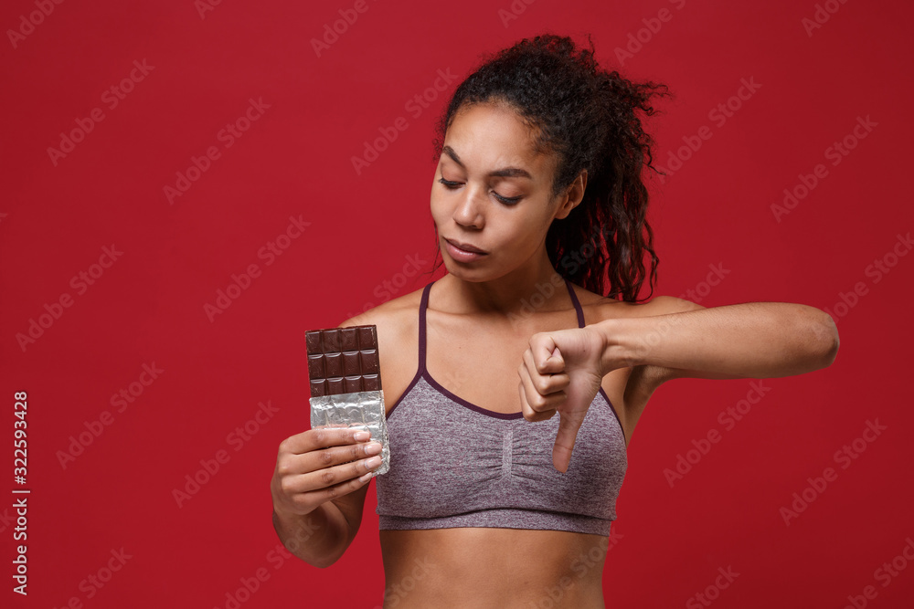 Young african american sports fitness woman in sportswear working out isolated on red background studio portrait. Sport exercises healthy lifestyle concept. Hold chocolate bar, showing thumb down. Young african american sports fitness woman in sportswear working out isolated on red background studio portrait. Sport exercises healthy lifestyle concept. Hold chocolate bar, showing thumb down.