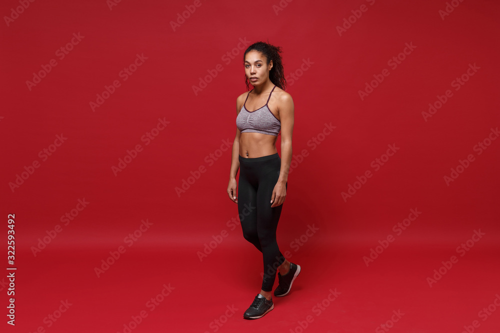 Fototapeta premium Beautiful young african american sports fitness woman in sportswear posing working out isolated on bright red background studio portrait. Sport exercises healthy lifestyle concept. Looking camera.