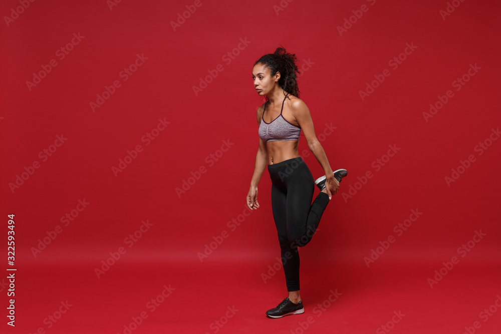Beautiful young african american sports fitness woman in sportswear posing working out isolated on red wall background. Sport exercises healthy lifestyle concept. Looking aside, stretching her legs.
