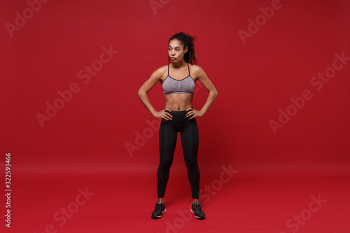 Strong young african american sports fitness woman in sportswear working out isolated on red background. Sport exercises healthy lifestyle concept. Standing with arms akimbo on waist, looking aside. © ViDi Studio