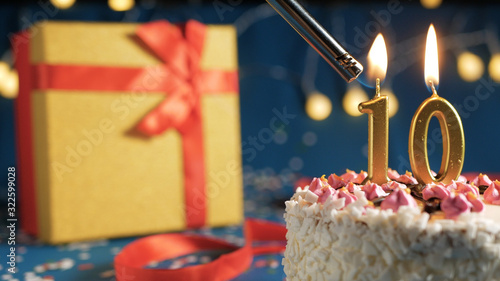 White birthday cake number 10 golden candles burning by lighter, blue background with lights and gift yellow box tied up with red ribbon. Close-up