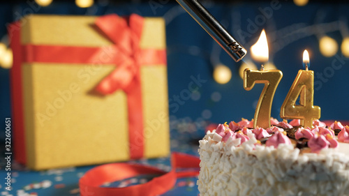 White birthday cake number 74 golden candles burning by lighter, blue background with lights and gift yellow box tied up with red ribbon. Close-up