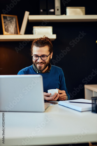 Young smiling handsome caucasian freelancer sitting in his modern home office and holding cup of coffee.