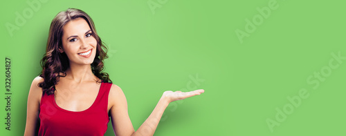 Happy smiling woman giving, holding or showing something or product or copy space for some text or slogan, green background. Brunette model in casual red clothing posing at studio.