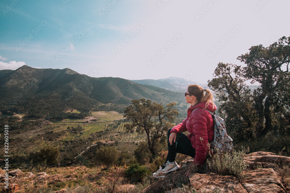 Fototapeta premium Young woman enjoying view on mountains in sunset, beautiful nature scenery with mountain tops. Girl or woman traveler with backpack alone, woman travelling solo, view from behind, vacation concept