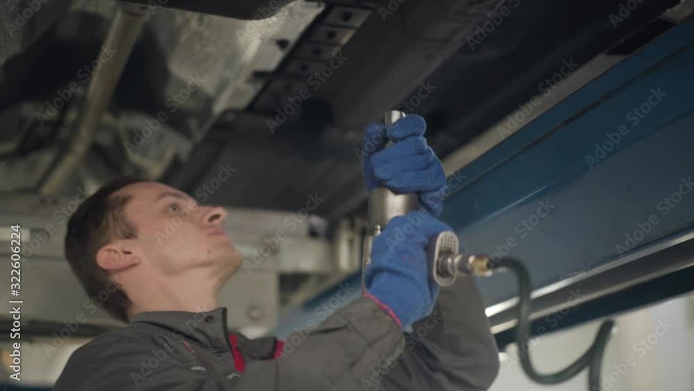 Close-up side view of confident Caucasian man in workwear using professional equipment for car maintenance. Young male auto mechanic fixing vehicle failure. Service station, repair shop. Close-up side view of confident Caucasian man in workwear using professional equipment for car maintenance. Young male auto mechanic fixing vehicle failure. Service station, repair shop.