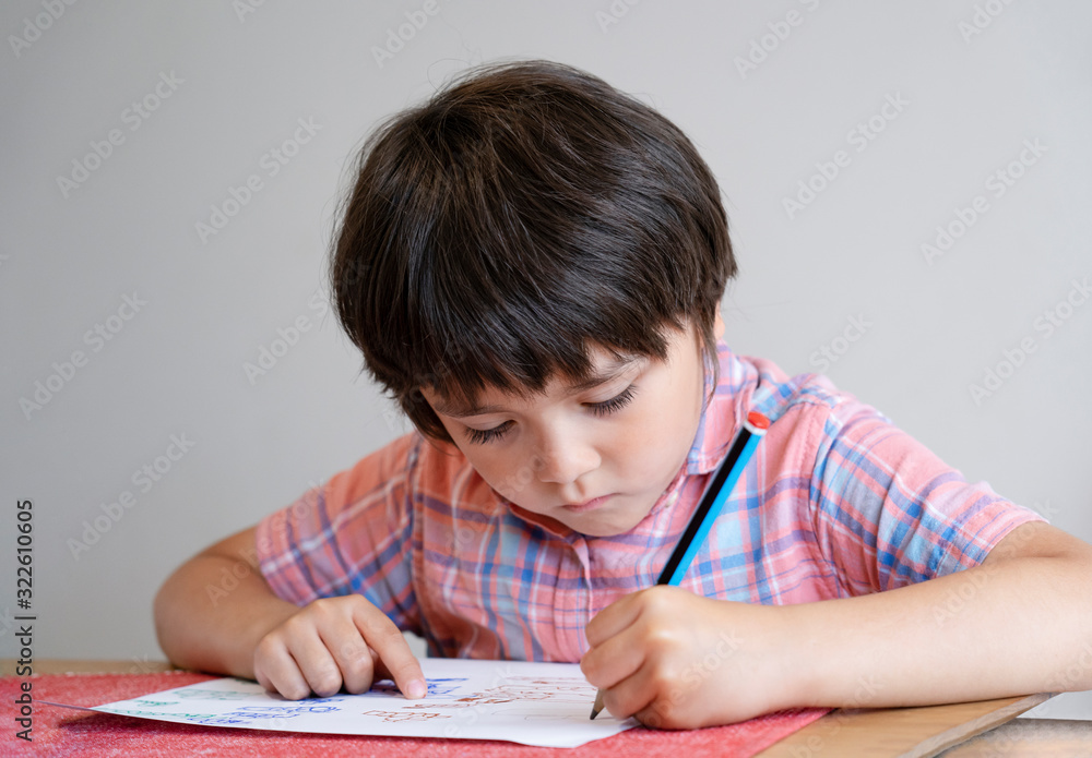 Portrait of school kid boy siting on table doing homework, Happy Child holding pencil writing, A boy drawing on white paper at the table,Elementary school and homeschooling concept Portrait of school kid boy siting on table doing homework, Happy Child holding pencil writing, A boy drawing on white paper at the table,Elementary school and homeschooling concept