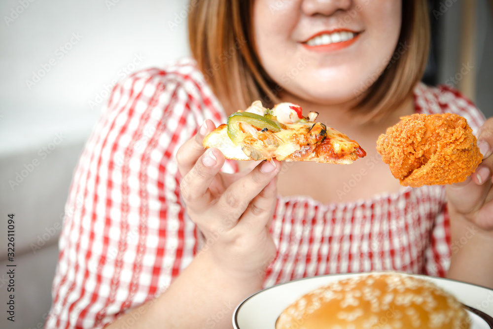 An Asian fat woman is eating pizza and fried chicken on the sofa in her house. The concept of consuming food that causes disease is not good for health An Asian fat woman is eating pizza and fried chicken on the sofa in her house. The concept of consuming food that causes disease is not good for health