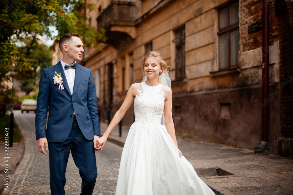 Wedding photo shooting. Bride and bridegroom walking in the city. Married couple embracing and looking at each other. Holding bouquet. Outdoor, full body Wedding photo shooting. Bride and bridegroom walking in the city. Married couple embracing and looking at each other. Holding bouquet. Outdoor, full body