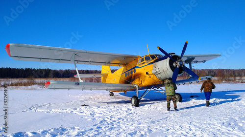 Fototapeta Naklejka Na Ścianę i Meble -  A yellow old biplane plane is parked on a winter airfield with technicians at work against a bright blue sky and white snow with the engine running and smoke