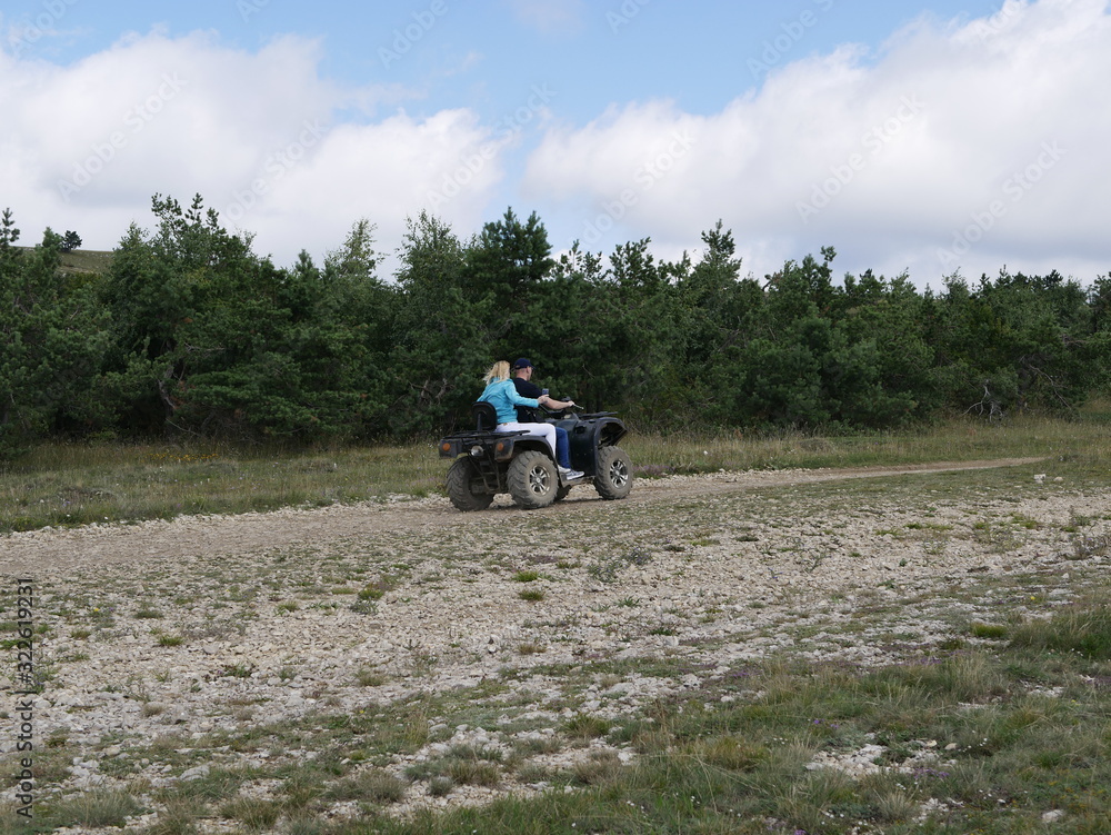Obraz premium Young man and woman people riding without protective gear on a black Quad bike on a background of forest and blue sky with clouds on a Sunny summer day