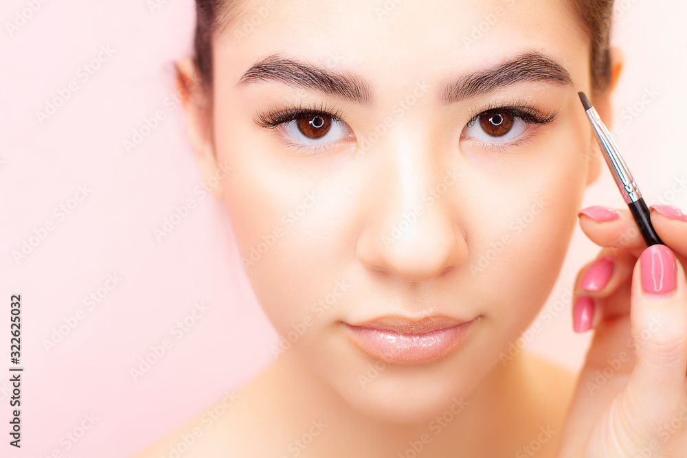 Eyebrow correction close up. A young woman of Asian appearance makes eyebrow correction on a pink background. Woman portrait on a pink background.