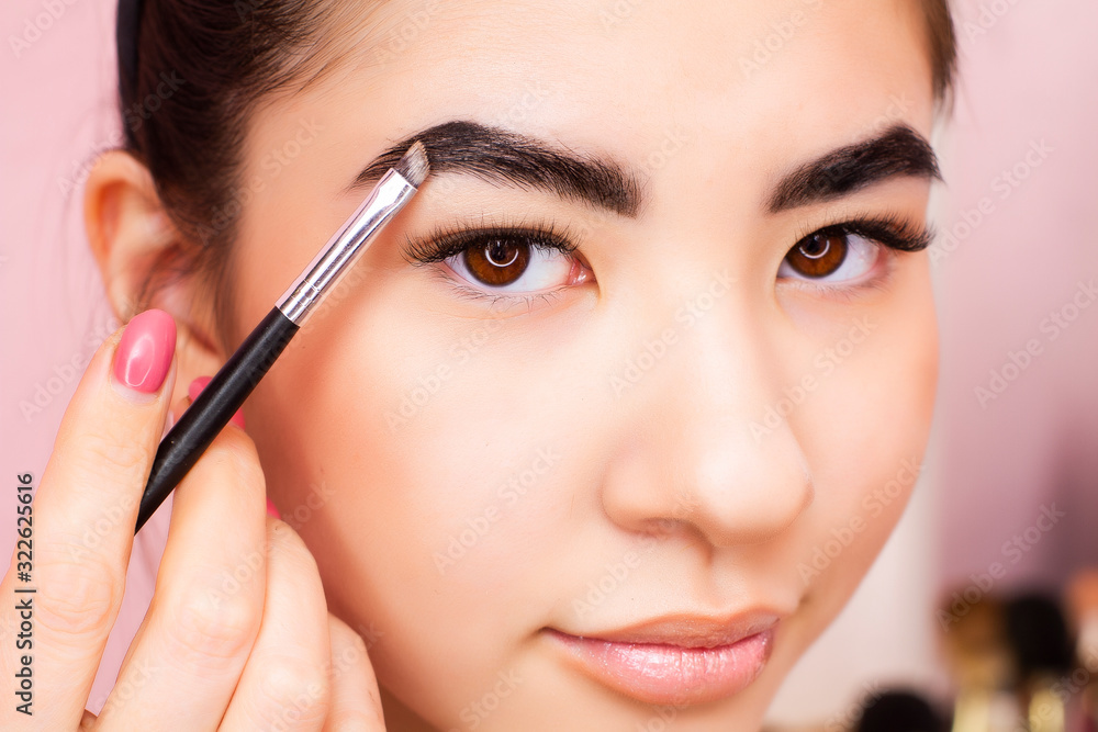 Eyebrow tinting procedure. Young brunette woman colors eyebrows in a beauty salon. Close-up model on a pink background.