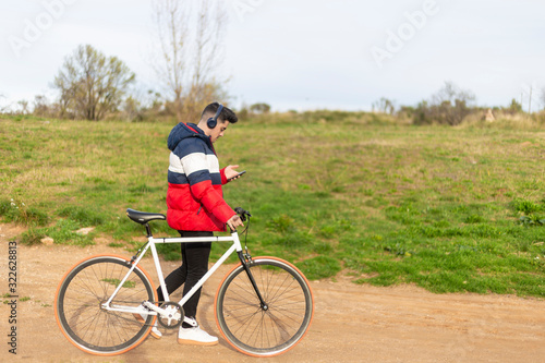 Handsome hipster young man enjoying a bicycle ride and using a smartphone while walking on park