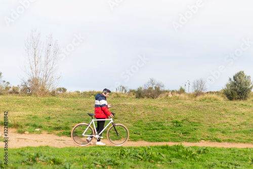 Handsome hipster young man enjoying a bicycle ride and using a smartphone while walking on park