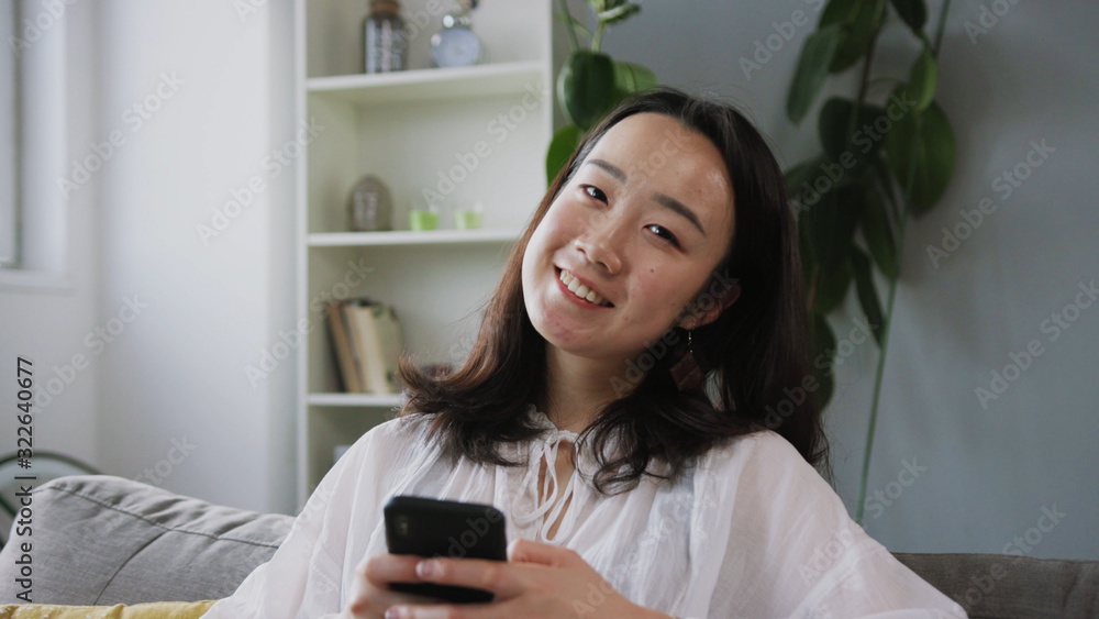 Close-up shooting of good-looking young Chinese woman in business clothes sitting on sofa and using smartphone. Fascinating girl looking at camera. Friendly face. Smile. Apartment. Close-up shooting of good-looking young Chinese woman in business clothes sitting on sofa and using smartphone. Fascinating girl looking at camera. Friendly face. Smile. Apartment.